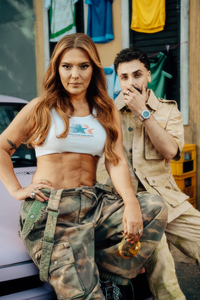 Trendsetting duo in urban attire posing outdoors with car and colorful backdrop.